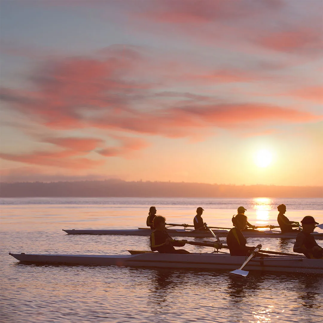 Persone che remano su barche sottili sull'acqua al tramonto con cielo arancione e rosa.