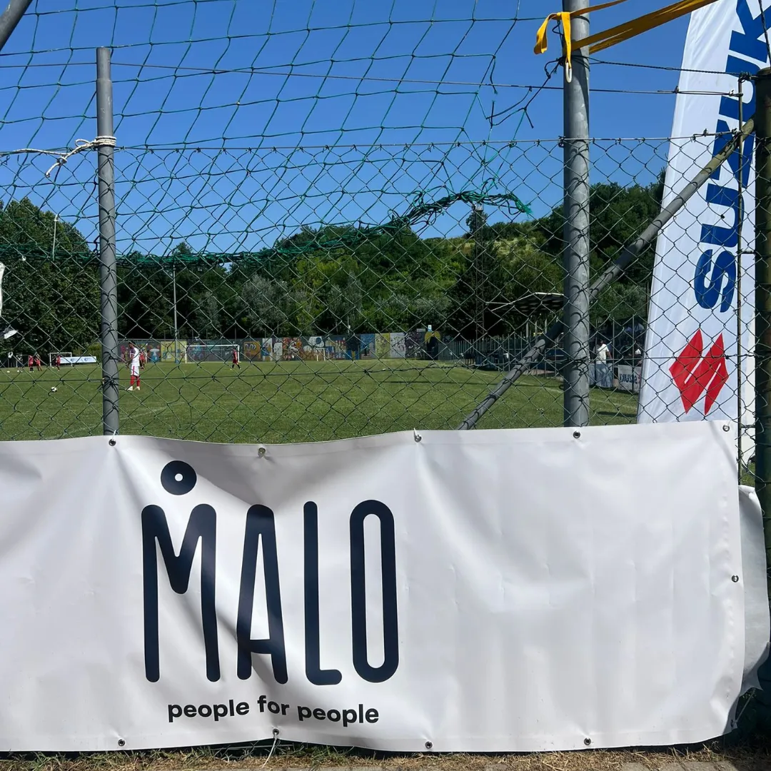 Campo da calcio con giocatori in divisa rossa visibile attraverso una rete, con uno striscione bianco con la scritta 'MALO people for people' in primo piano.