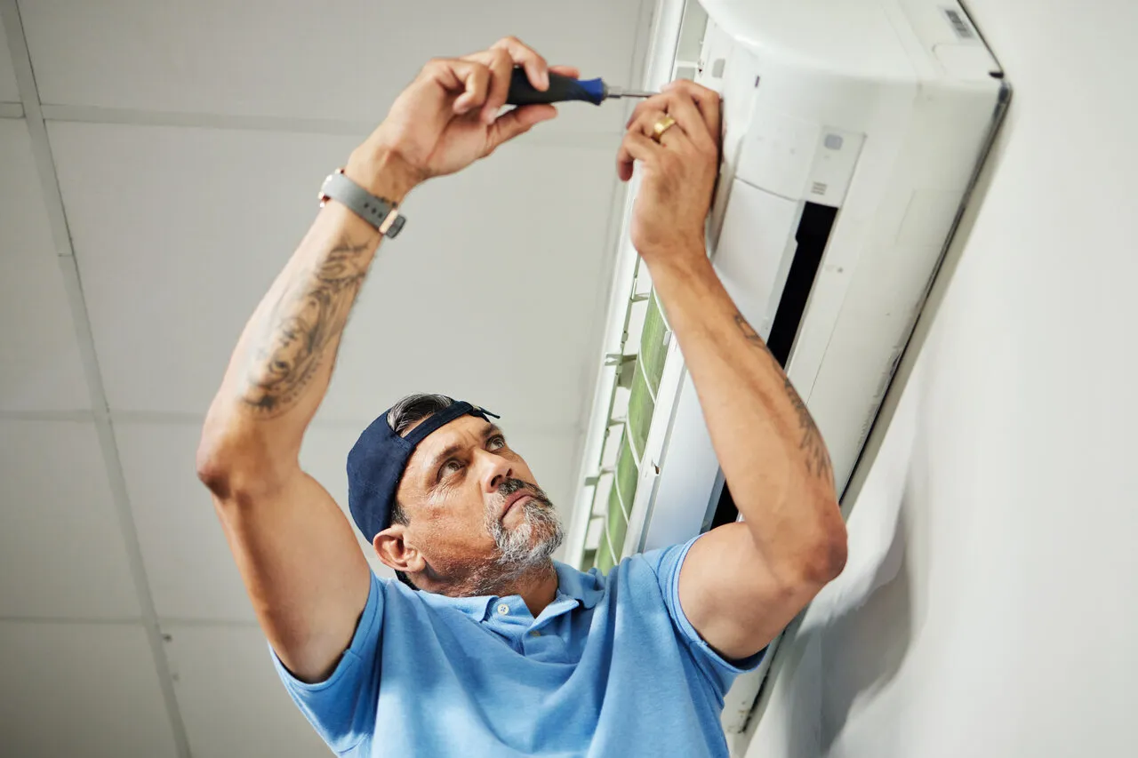 Man in a blue polo and cap repairs an air conditioner on a white wall.