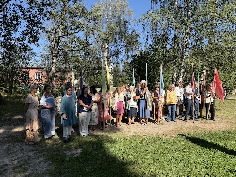 Grupp av människor stående utomhus i en park med flaggor under en solig dag.