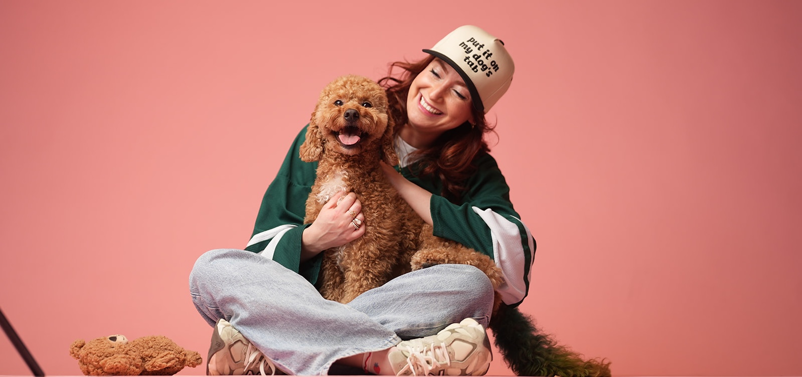 Smiling woman in a cap hugs a happy curly brown dog seated on her lap against a pink background.