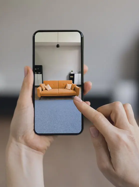 Eine Person nutzt eine Augmented-Reality-App auf dem Smartphone, um ein orangefarbenes Sofa virtuell im eigenen Raum zu platzieren. augmented-reality-ar-app-3d-visualisierung-virtuelle-anprobe-möbel-amazon-agentur-visual-conversion
