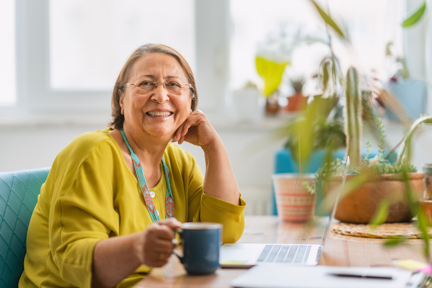 A Old lady sitting holding a cup of tea with her laptop open on desk smiling towards the camera.
