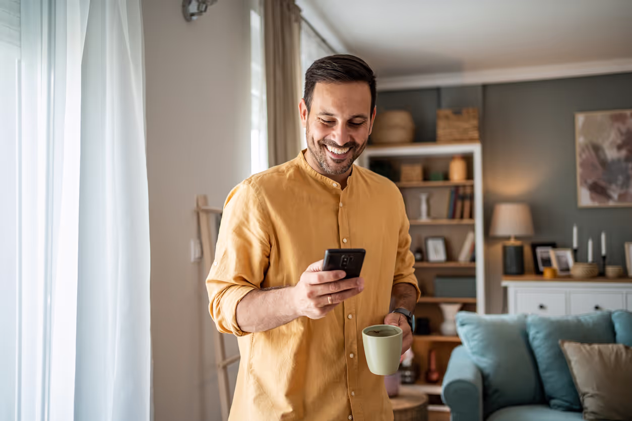 A man in orange short looking at his phone and smiling while having a cup in other hand