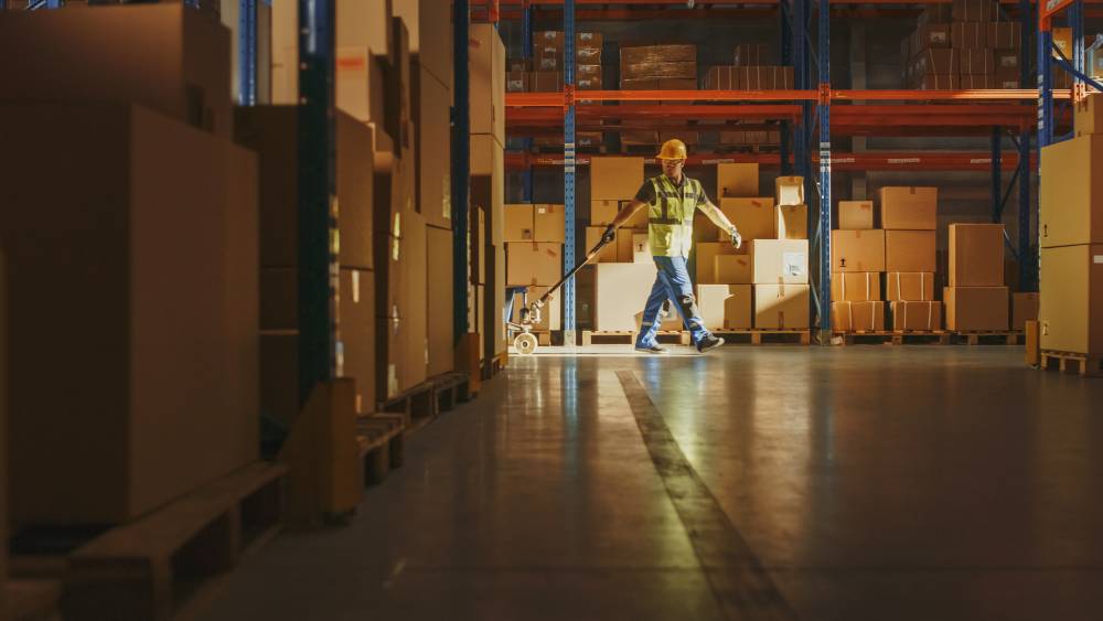 Warehouse worker pulling a pallet jack past shelves of boxes.