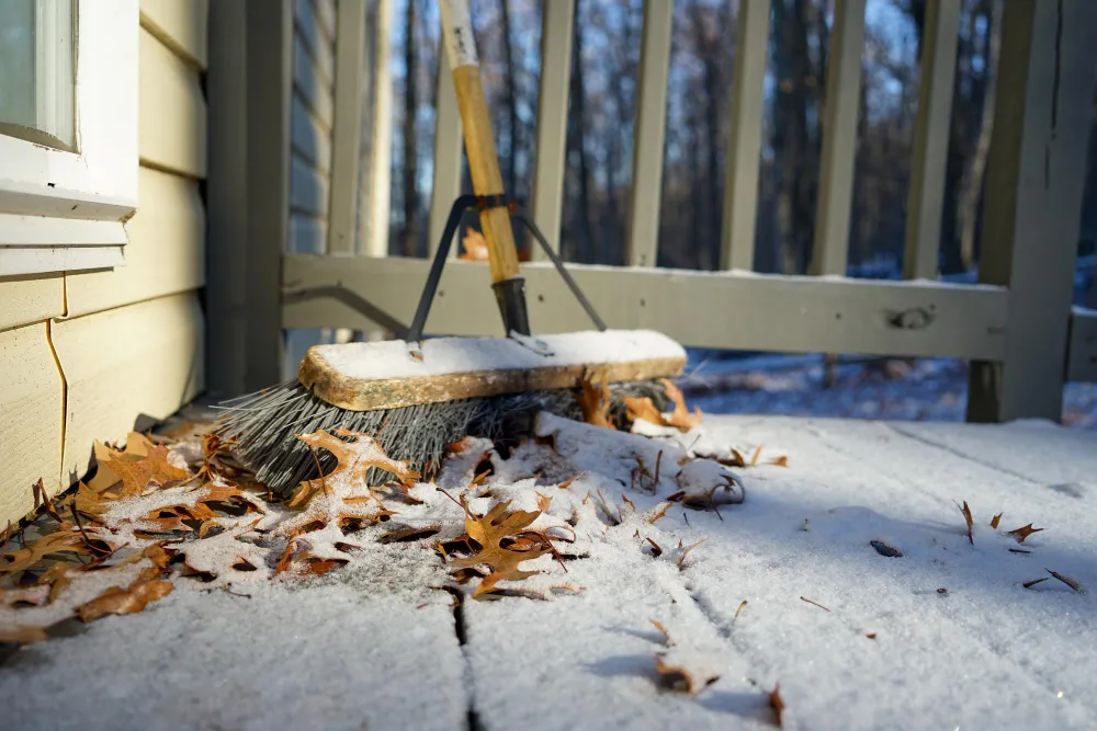 Besen und Herbstblätter auf verschneiter Veranda mit Holzzaun im Hintergrund