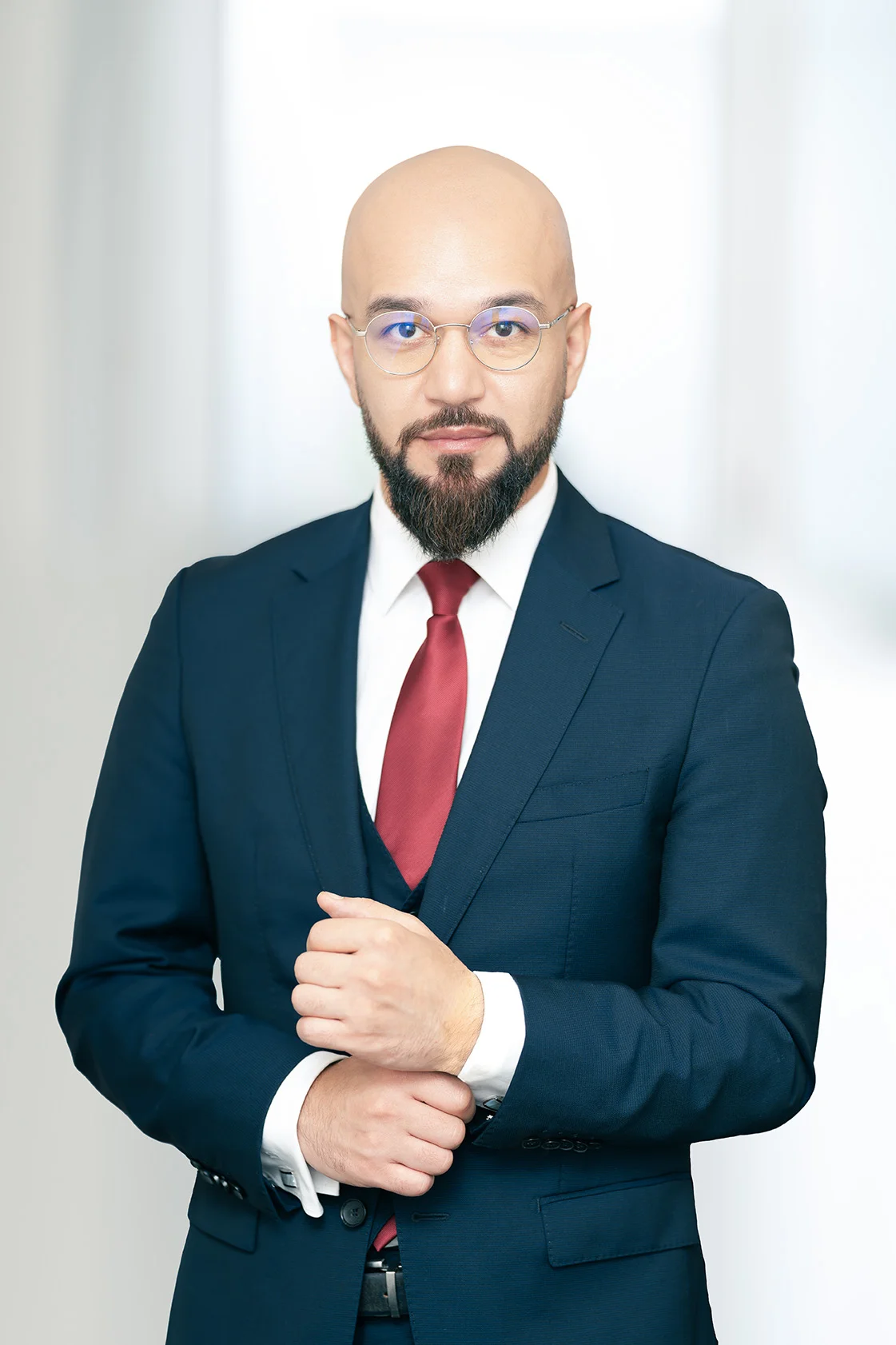 Porträt eines bärtigen Mannes mit Brille in blauem Anzug und roter Krawatte vor hellem Hintergrund.