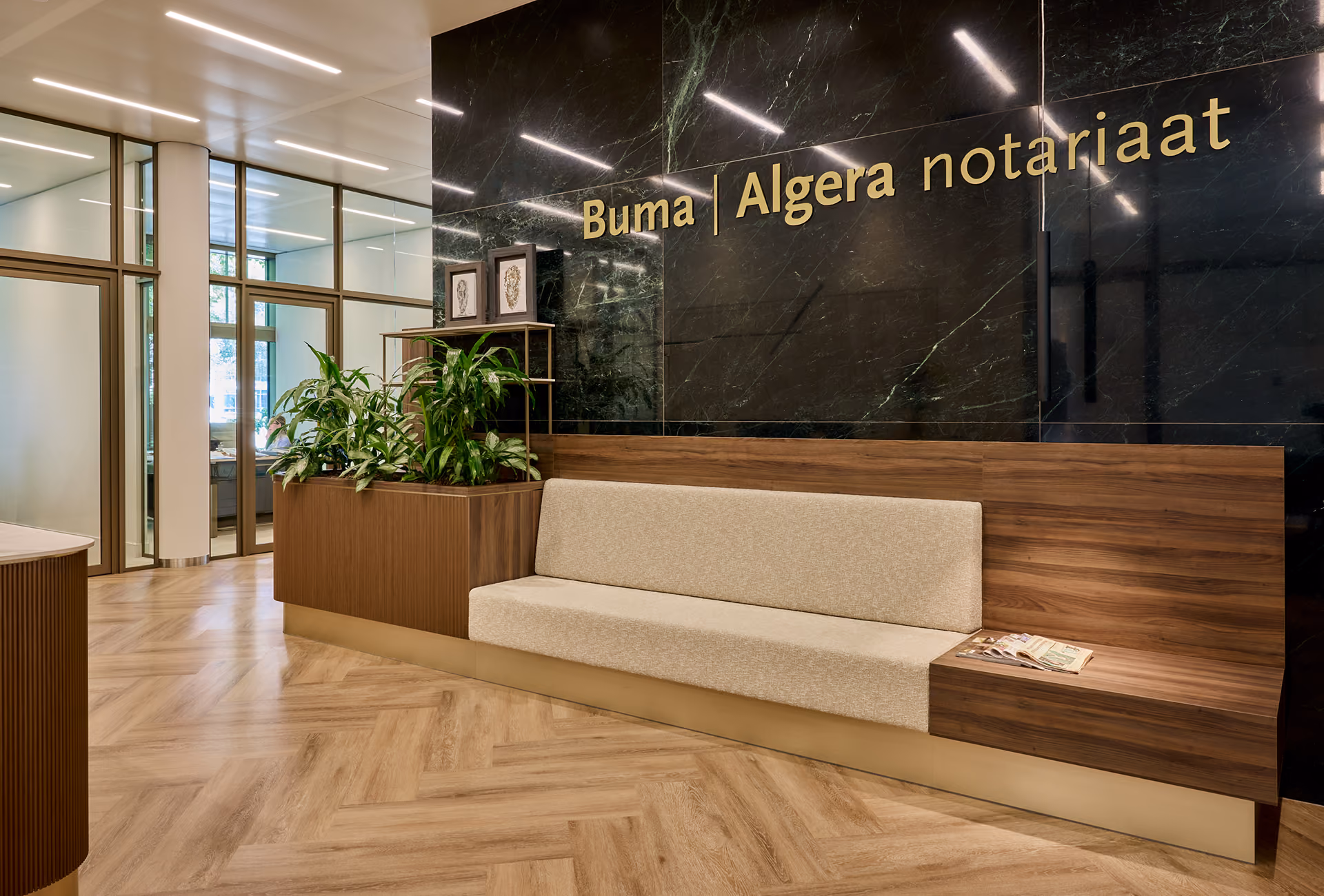Moderne kantoorruimte met houten vloer en groene planten, met een beige zitbank aan een houten wand en de tekst 'Buma | Algera notariaat' in gouden le