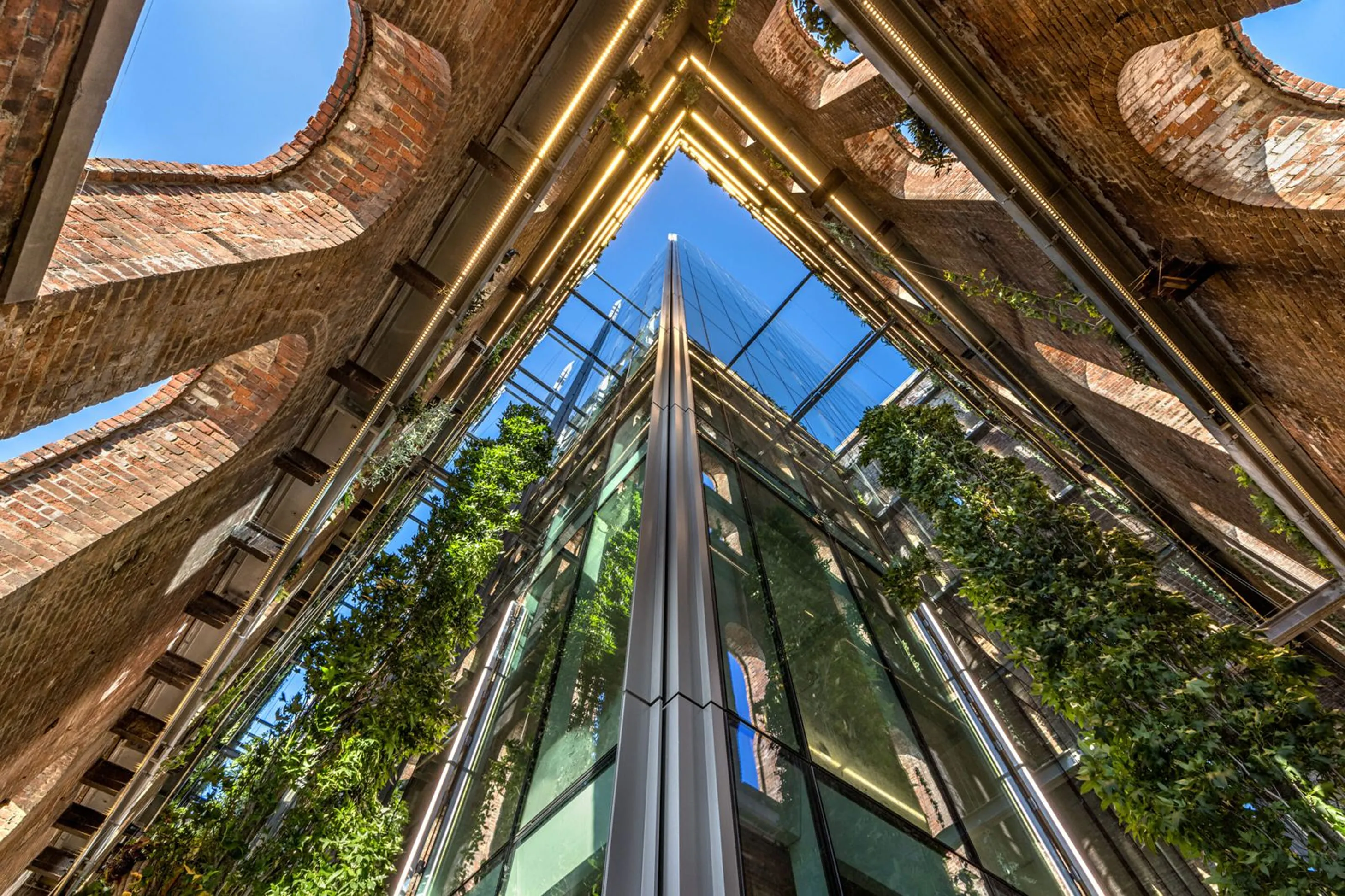 Upward view of a modern glass building facade surrounded by old brick walls with climbing green plants and integrated lighting.