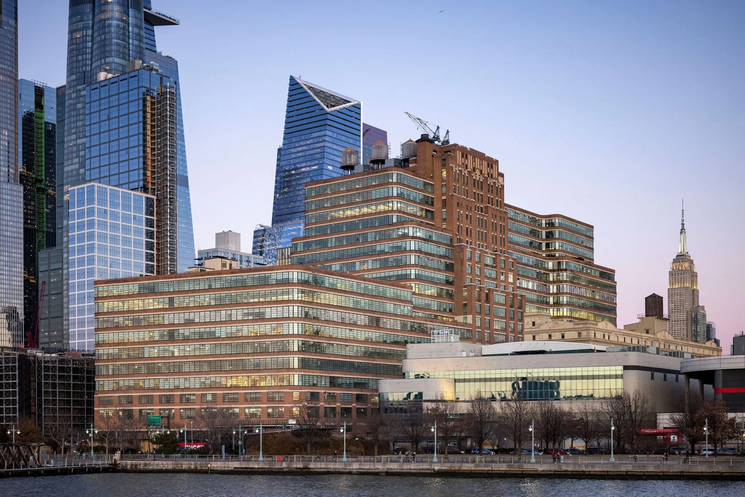 City skyline featuring modern glass skyscrapers and a large brick building by the waterfront at dusk, with the Empire State Building visible in the background.