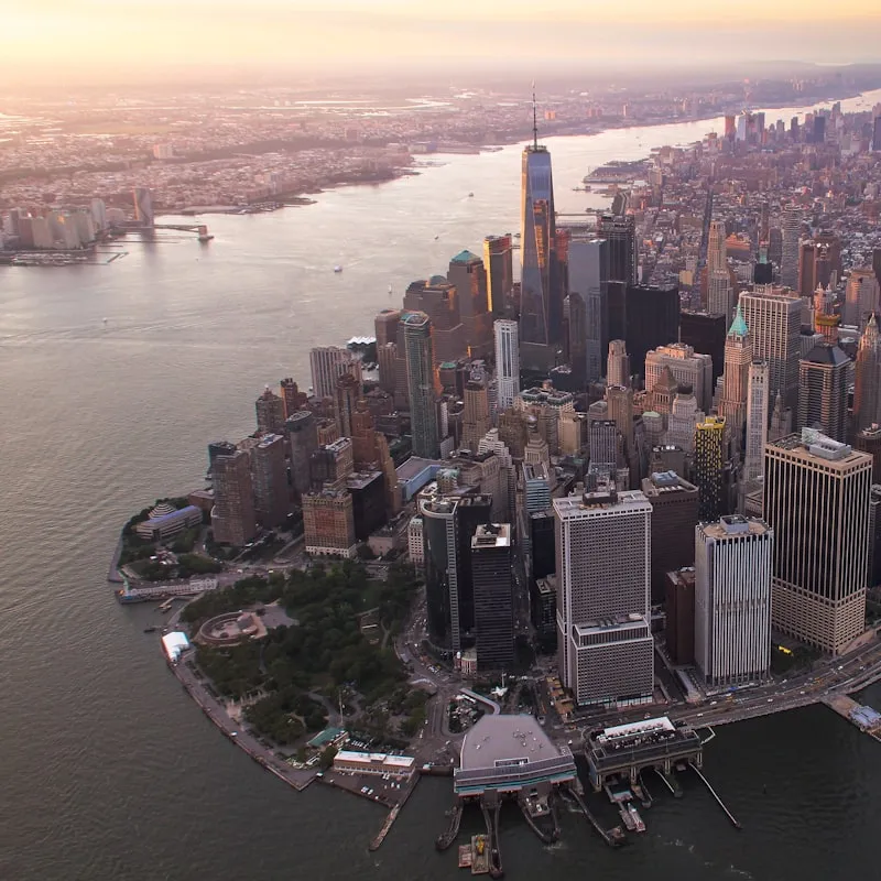 Aerial view of lower Manhattan skyline at sunset, featuring One World Trade Center and surrounding skyscrapers by the waterfront.