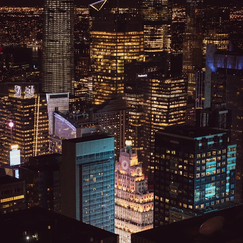 Nighttime cityscape of illuminated skyscrapers with a prominent clock tower building in the center.