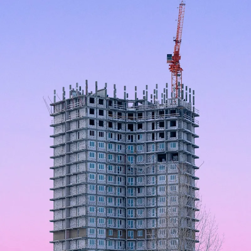 Under-construction tall concrete building with multiple floors and a red construction crane on top against a clear sky.