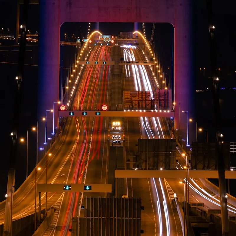 Night view of a lit bridge with multiple lanes showing streaks of red and white light from moving vehicles.