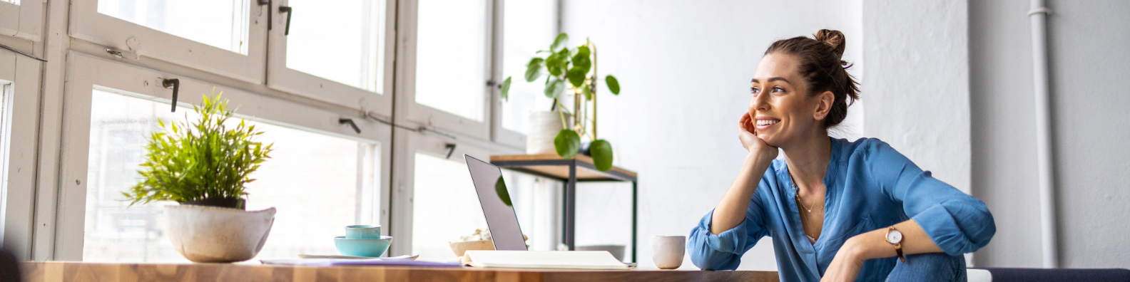Smiling woman in blue top working at home office with laptop and plants