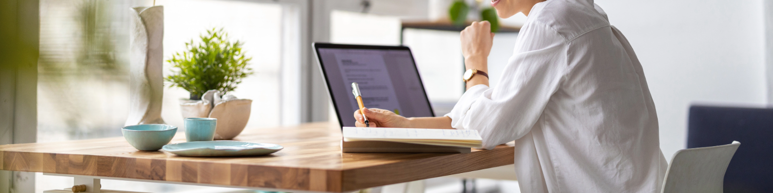 Person working at wooden desk with laptop, plant, and blue ceramic dishes