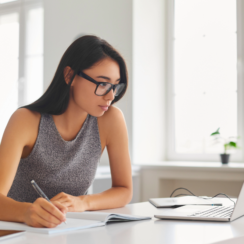 Woman wearing glasses studying and taking notes at laptop in bright room