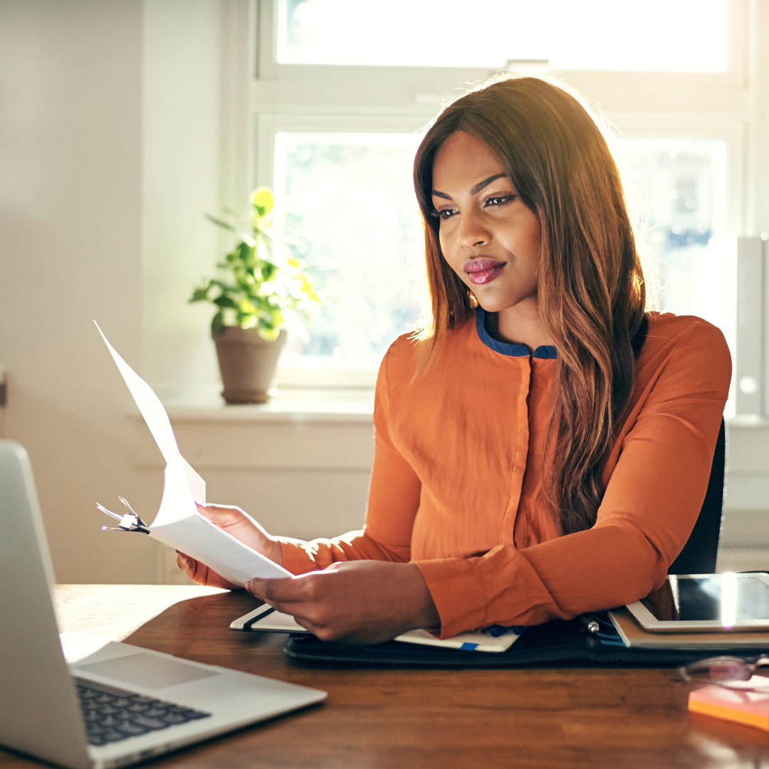 Woman in orange shirt working at desk with laptop and documents