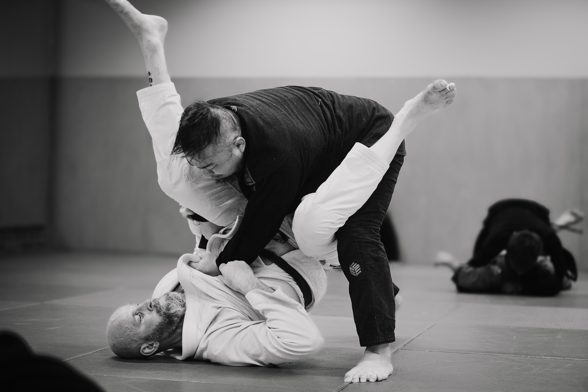 Two men practicing Brazilian Jiu-Jitsu on mats, with one man in a white gi on the ground and the other in a black gi standing over him.