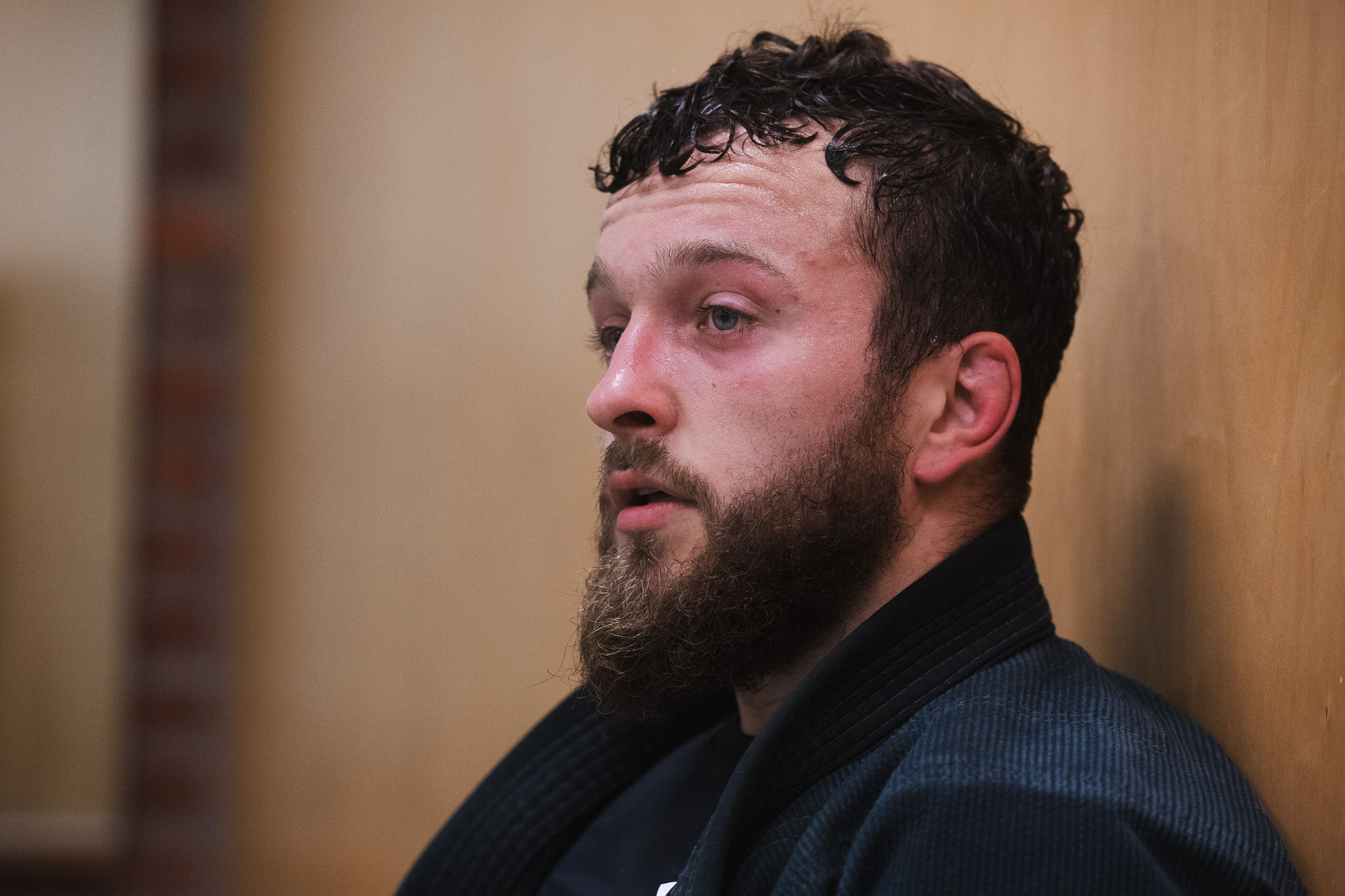 Man with a beard and wet hair wearing a dark martial arts gi sitting against a wooden wall looking thoughtful.