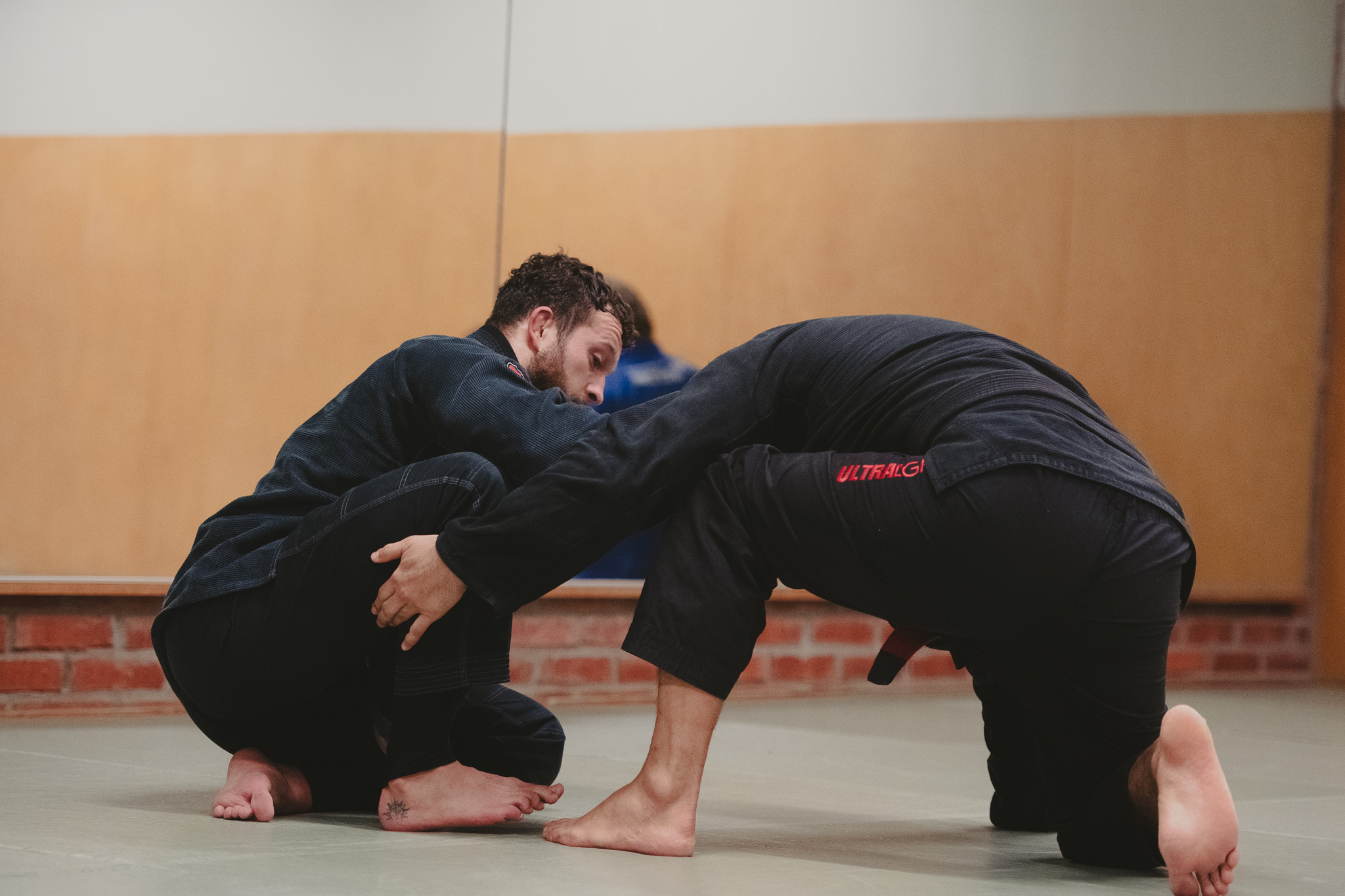 Two men wearing black martial arts gis practicing Brazilian Jiu-Jitsu on a mat in a gym.