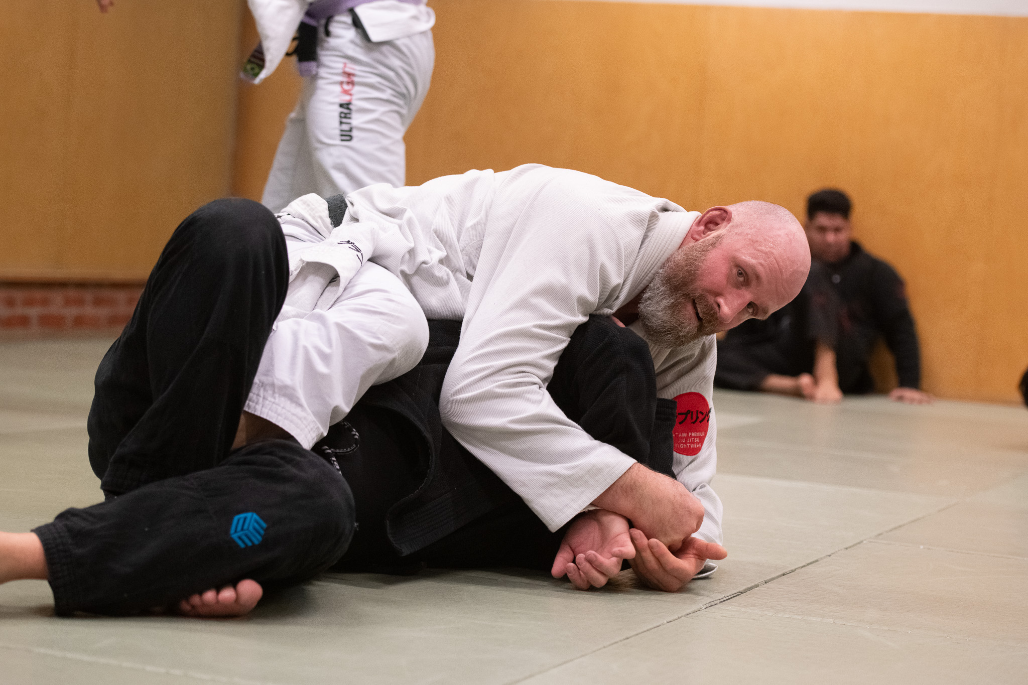 Two men practicing Brazilian Jiu-Jitsu grappling on mats inside a dojo, with one man securing a hold on the other.