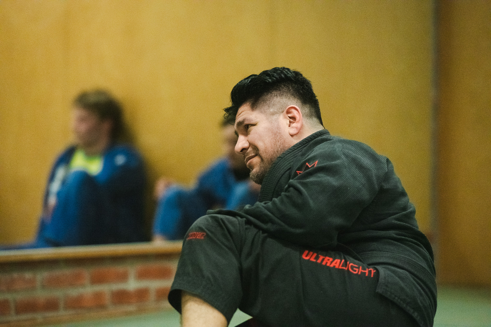 Man in a black martial arts gi with 'ULTRALIGHT' written on the pants, sitting and looking to the side with two blurred figures in the background.