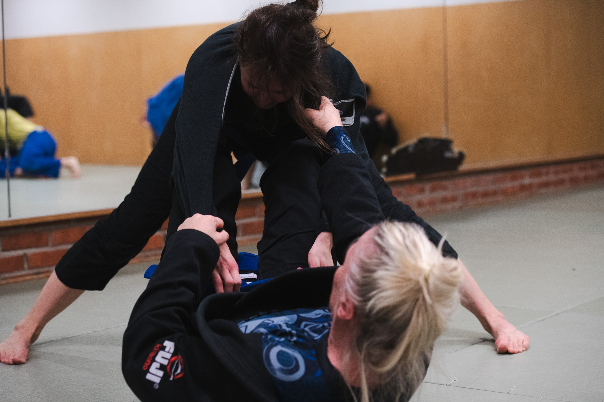Two women in black martial arts uniforms practicing Brazilian jiu-jitsu on mats in a dojo.