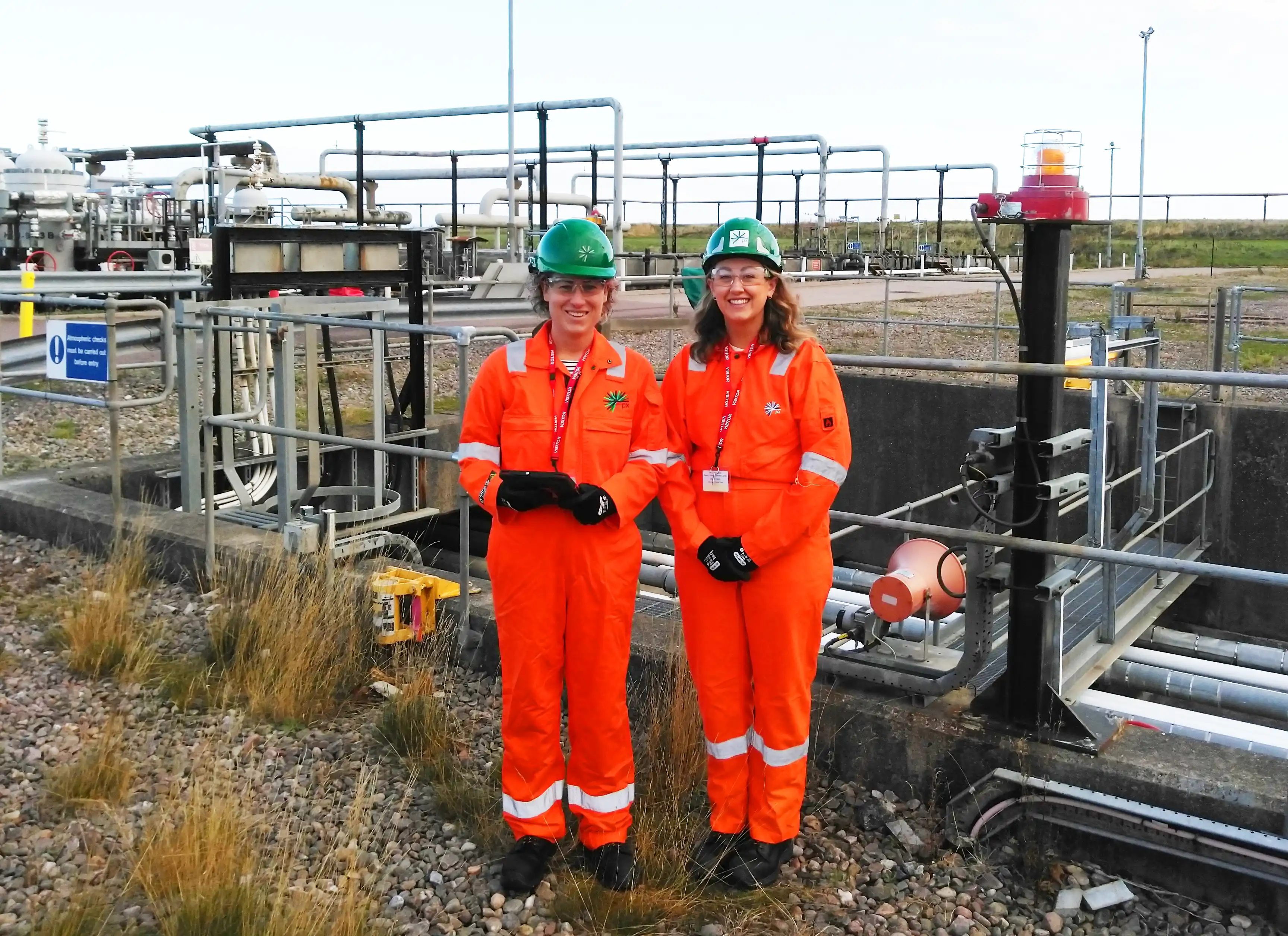 Claire Murray and Georgia Ledwidge from Arnlea wearing orange PPE and green hard hats, standing at PX Group’s St Fergus Gas Terminal site near Peterhead, surrounded by industrial equipment and pipework.
