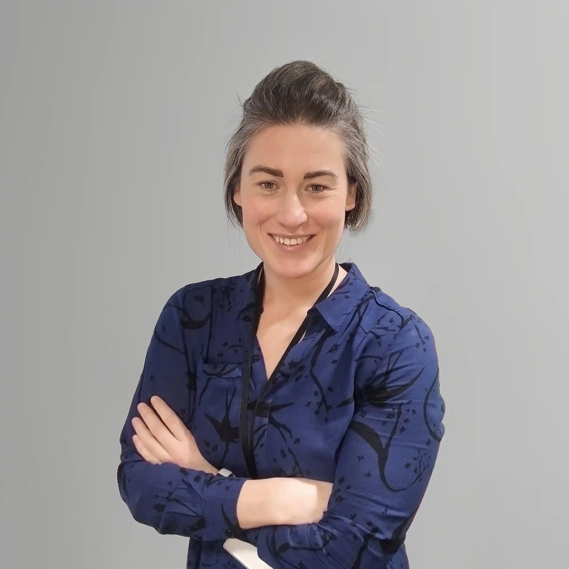“A woman standing with arms crossed, smiling, wearing a patterned dark blue shirt against a plain grey background.”