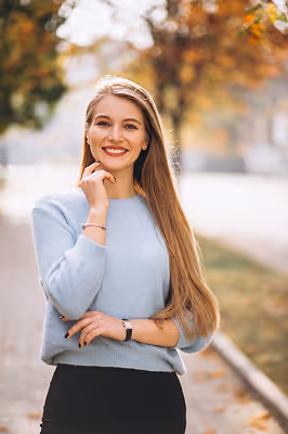 Smiling young woman with long blonde hair wearing a light blue sweater standing outdoors in a park during autumn.