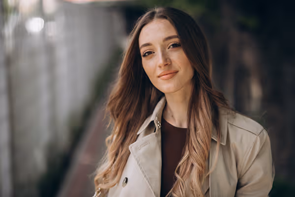 Smiling young woman with long light brown hair wearing a beige trench coat outdoors.