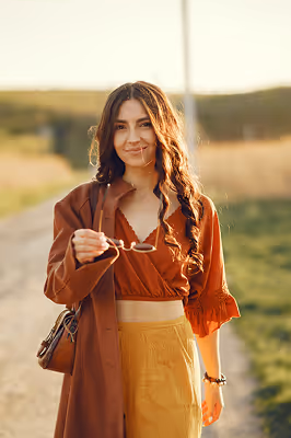Young woman with long curly hair wearing a rust-colored blouse and mustard pants, holding sunglasses while standing outdoors on a sunny day.