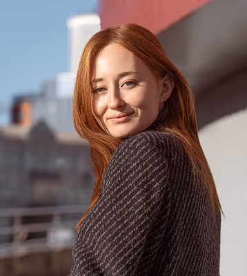 Red-haired woman wearing a dark textured sweater, smiling outdoors with blurred buildings in the background.