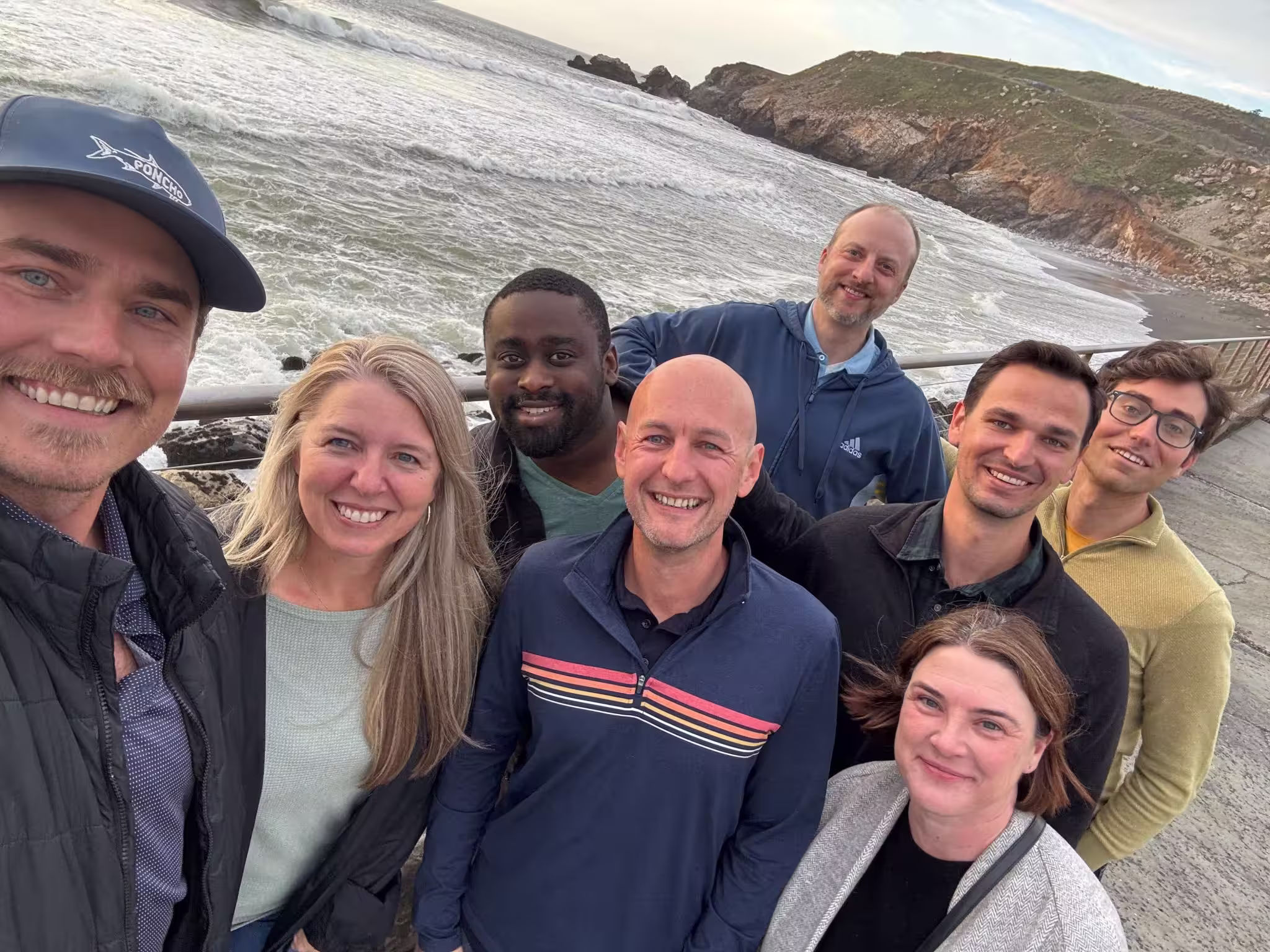Group of eight adults smiling and posing for a selfie near a rocky coastline with waves and hills in the background.