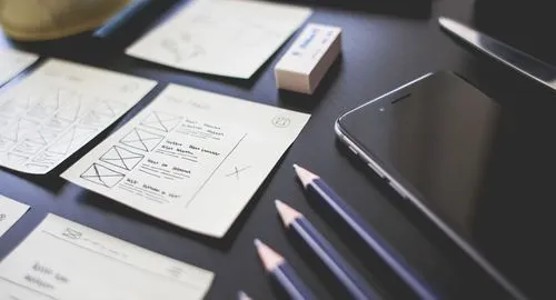 Desk with paper web design wireframe sketches, three blue pencils, an eraser, and a smartphone.