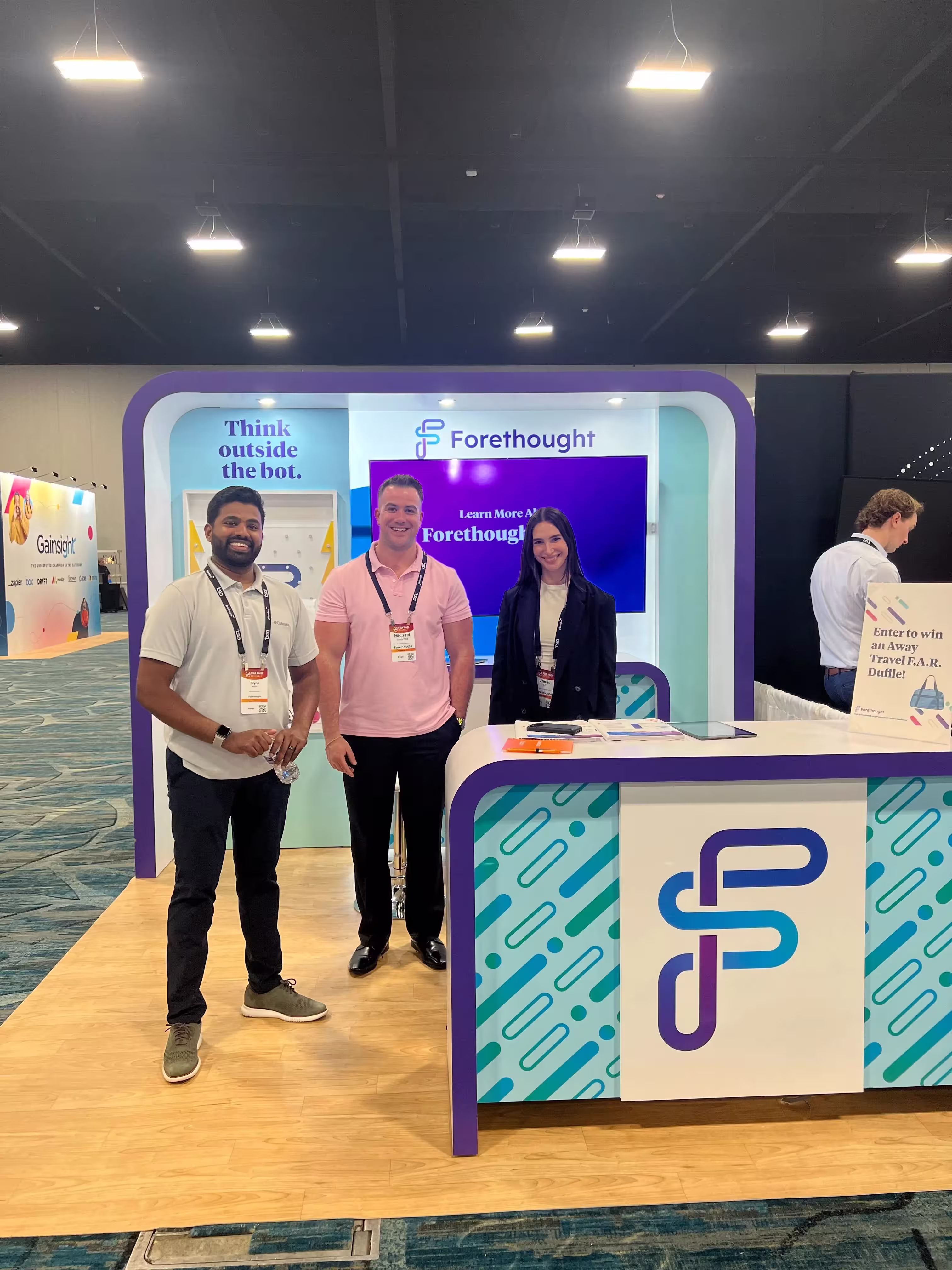 Three people smiling and standing in front of a Forethought booth at a conference with a sign that says 'Think outside the bot.'
