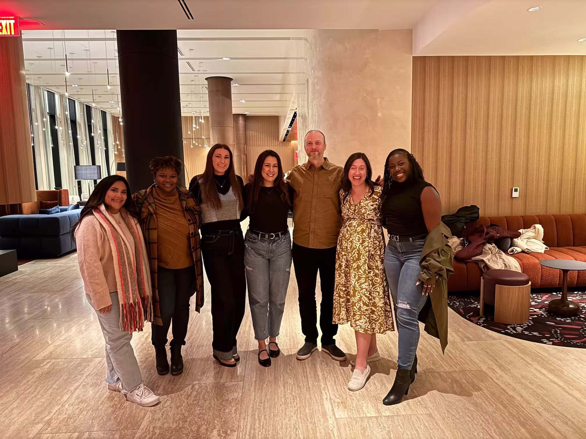 Group of six people standing closely together and smiling in a modern indoor setting with wooden walls and stylish furniture.
