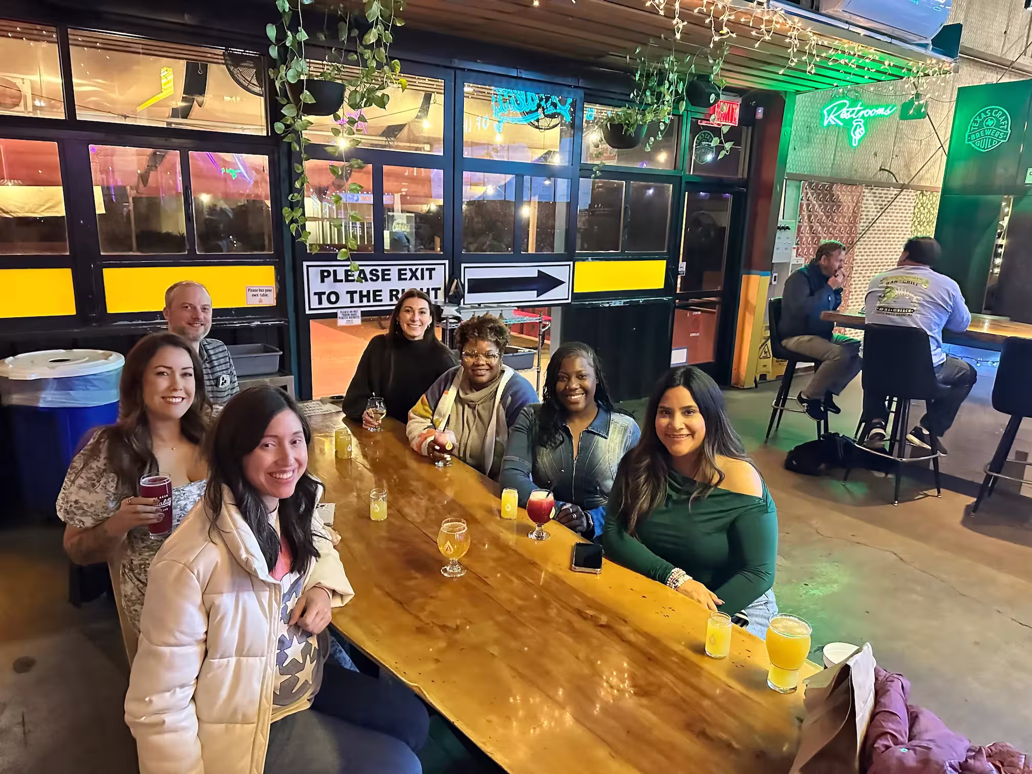 Group of seven people sitting around a wooden table with drinks in a brightly lit bar or brewery.