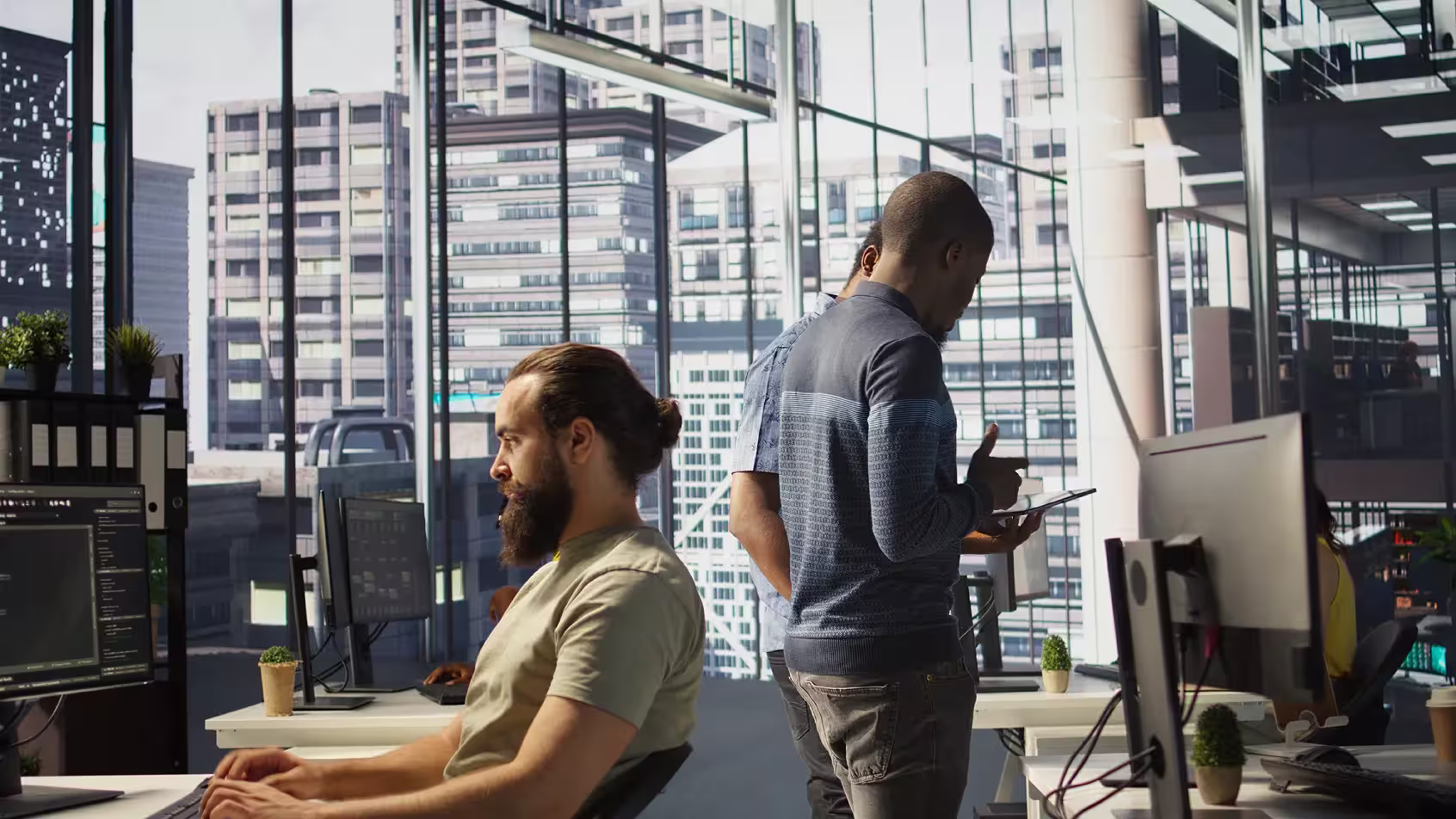 Office workers collaborating and working at computers in a modern workspace with large windows overlooking a cityscape.