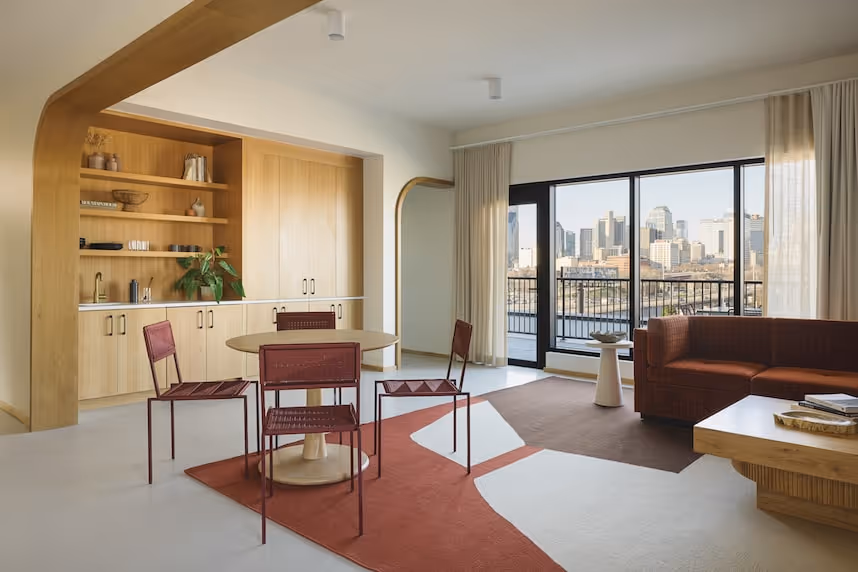 Open living space with pale floors, a red geometric rug, round dining table surrounded by four red metal chairs, built‑in wood cabinetry, and large windows overlooking a distant city skyline.