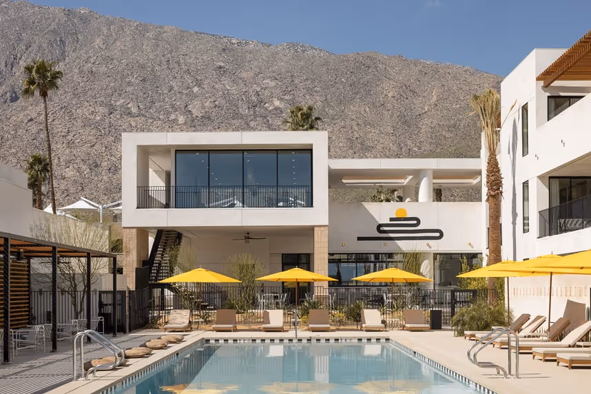 Rectangular swimming pool before a white two‑storey building with floor‑to‑ceiling windows, flanked by yellow umbrellas and lounge chairs, backed by rugged desert mountains.