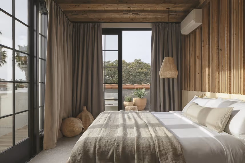 Cozy bedroom with a wood‑plank accent wall, neutral‑linen bed, woven pendant light, and floor‑to‑ceiling black‑framed windows opening to a balcony with potted plants and treetop views.