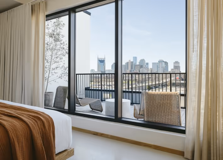Modern bedroom interior with floor-to-ceiling glass windows opening onto a balcony overlooking a distant city skyline