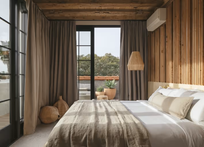 Cozy bedroom featuring wood-paneled accent wall, neutral linen bedding, rattan decor pieces, and glass doors leading to a balcony with potted plants