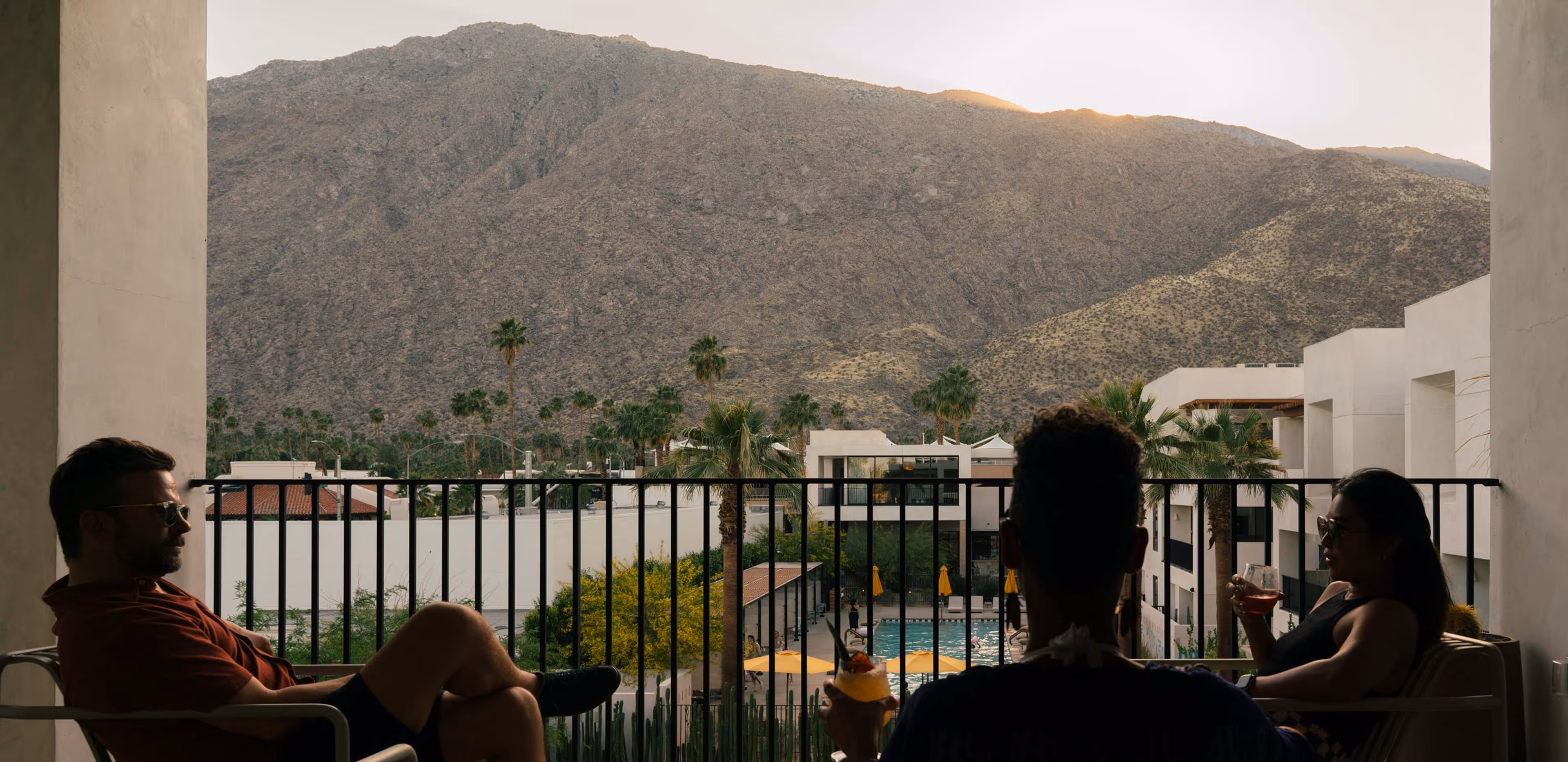 Three friends on a balcony at sunset, holding colorful cocktails and relaxing as they overlook a sparkling pool, palm trees, and a rugged mountain backdrop.