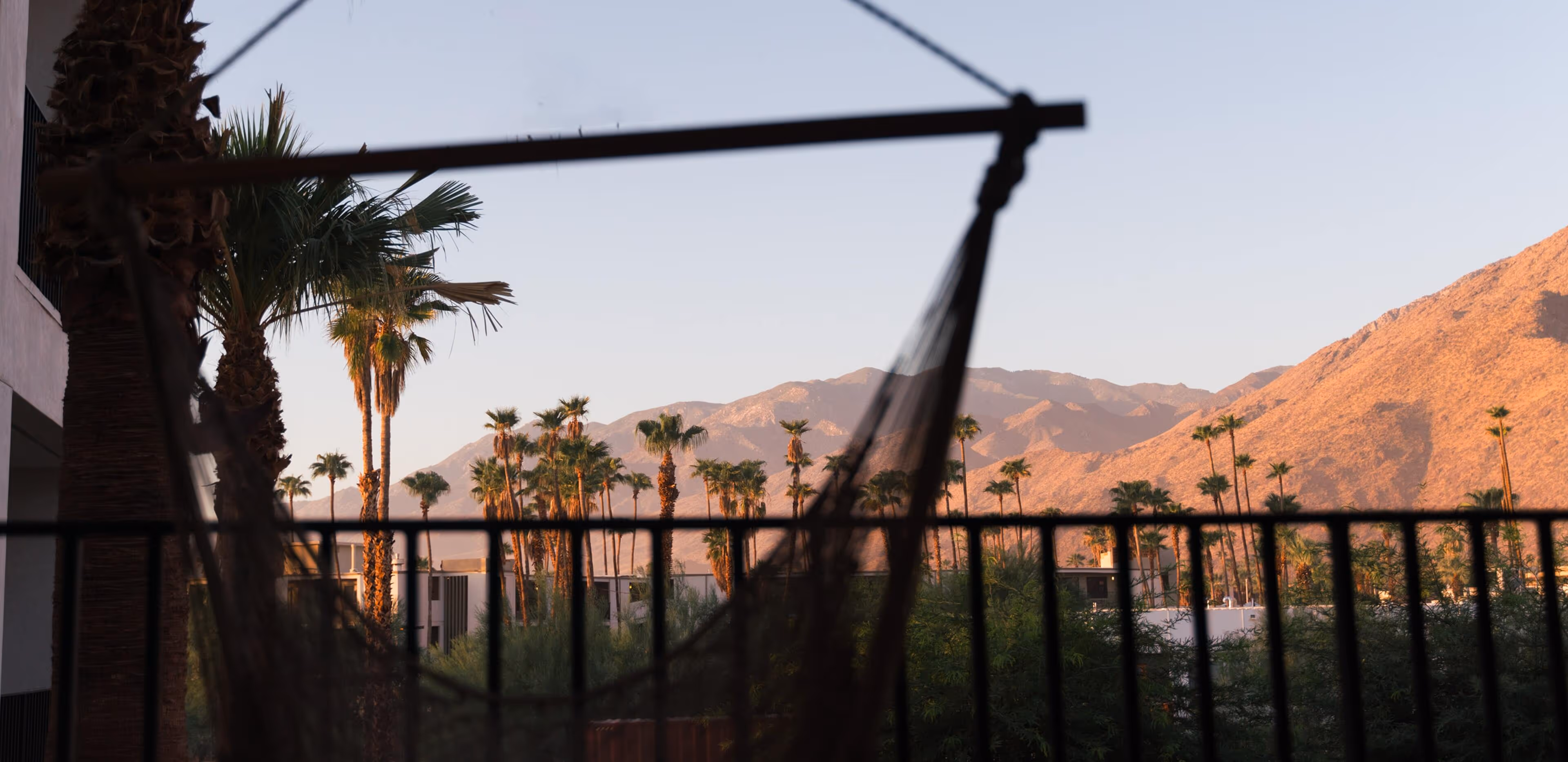 Silhouette of a hanging hammock on a balcony railing, overlooking palm trees and distant mountains bathed in golden sunset light.