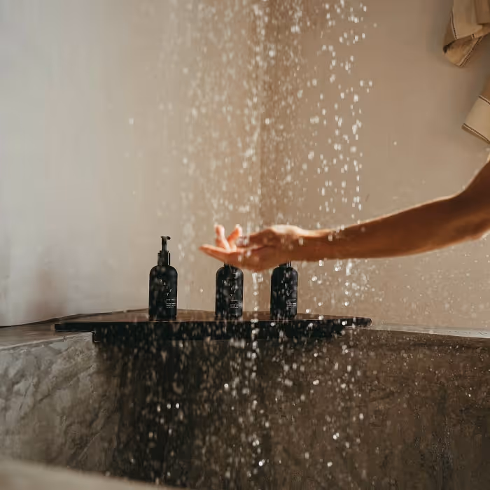 Hand catching falling water in a minimalist stone shower, with three black toiletries on a corner shelf and warm, natural light.