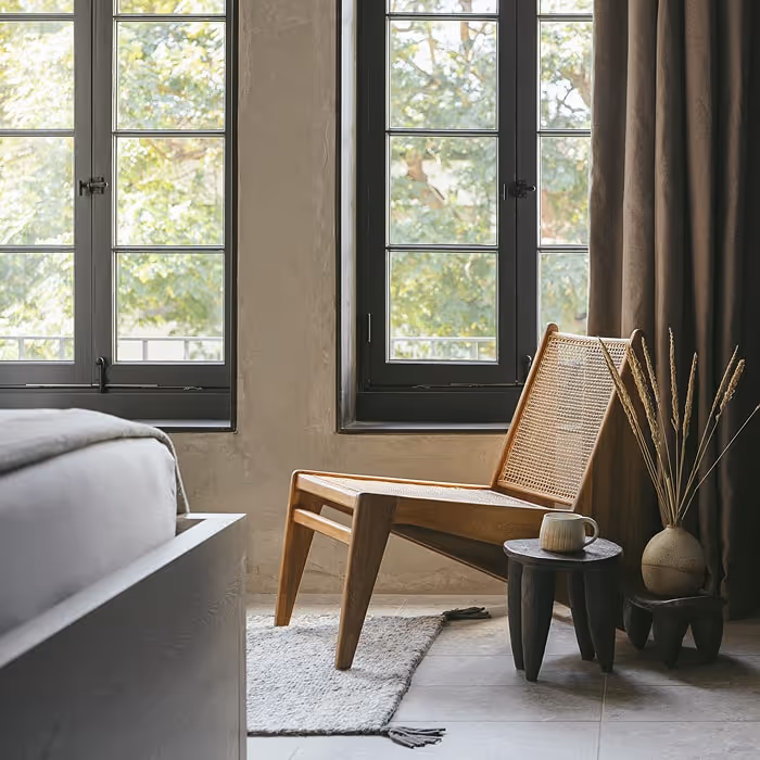 Minimalist bedroom corner with a woven chair and small side tables, set by large windows with sheer light. Decor includes a vase with dried grasses and a ceramic mug, creating a calm atmosphere.