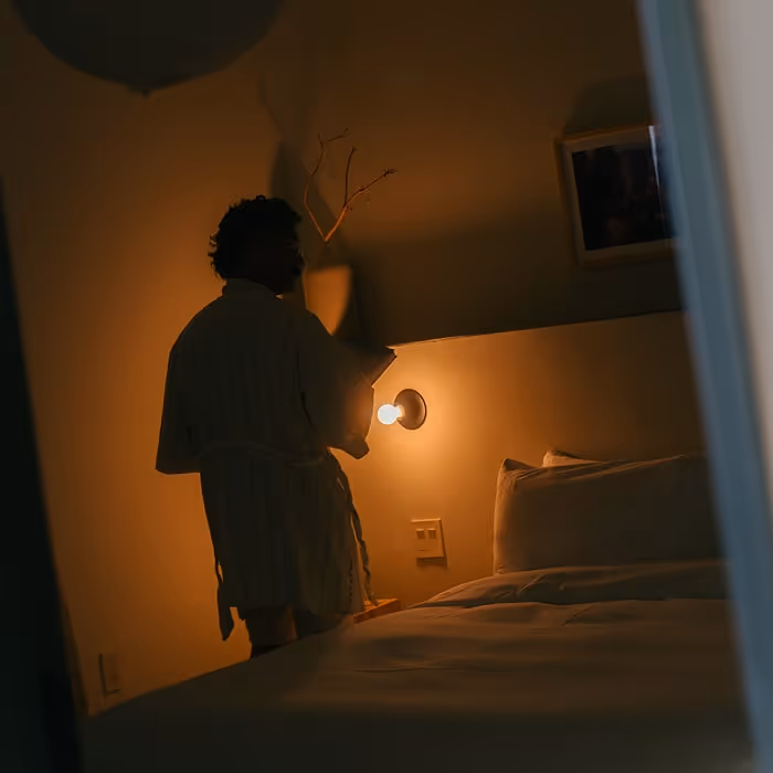 Person in a robe standing by a bedside lamp, reading a book in a dimly lit, cozy bedroom.